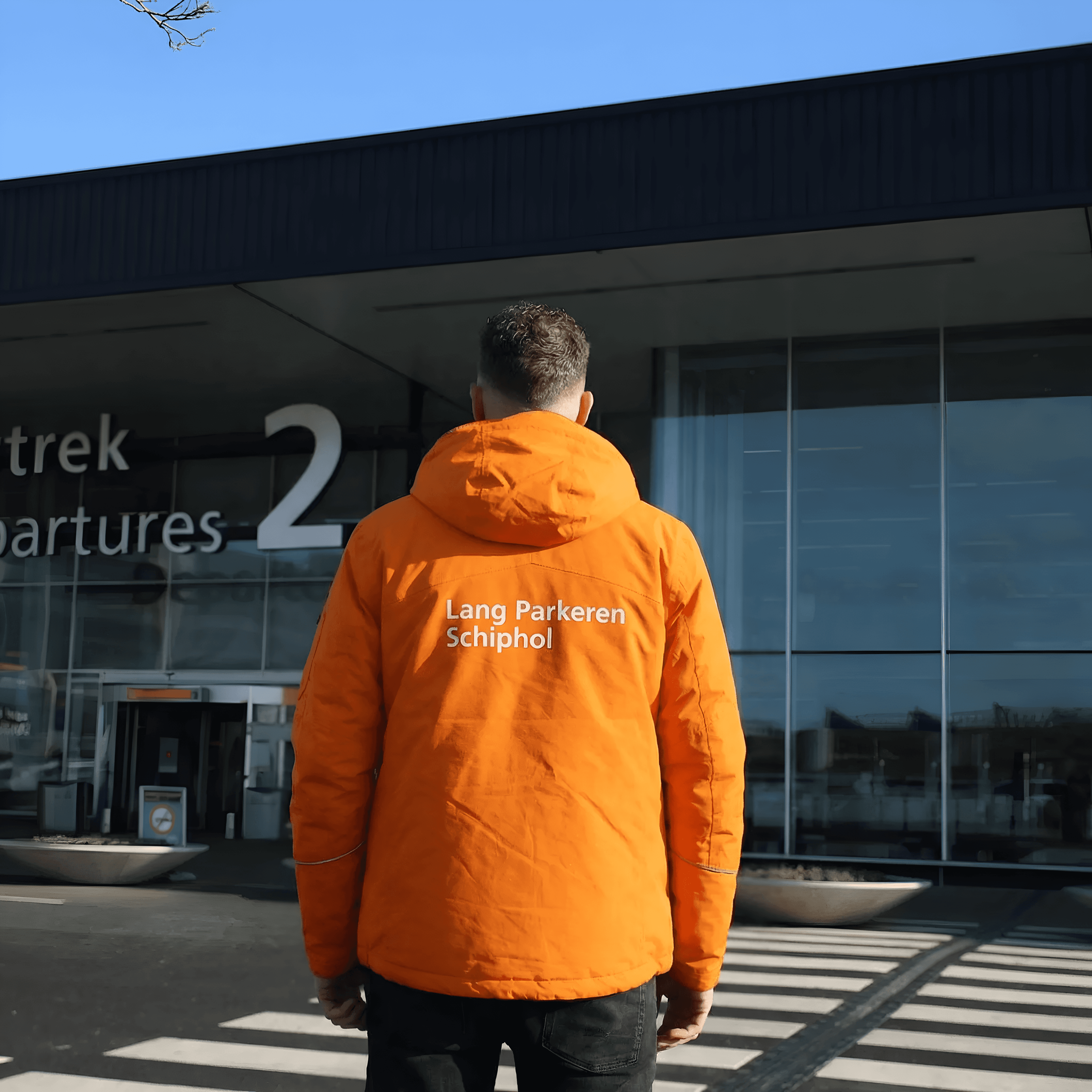 Chauffeur van Lang Parkeren Schiphol in oranje bedrijfsjas voor de vertrekhal Vertrek 2 op Schiphol.
